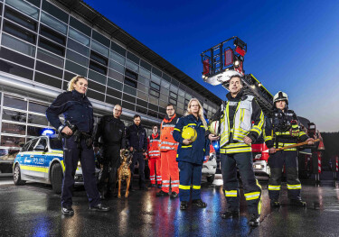 Mitarbeiterinnen und Mitarbeiter von Polizei, Rettungsdienst und Feuerwehr bei Dämmerung mit Fahrzeugen im Hintergrund.