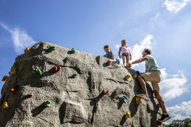 Vater mit zwei Kindern klettert bei Sonnenschein auf dem Boulderfelsen.